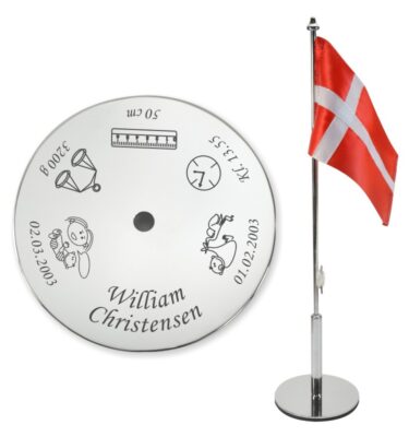 Navngivningsflag i poleret stål med personlig gravering af navn og fødselsdata, vist ved siden af det håndsyede danske flag.