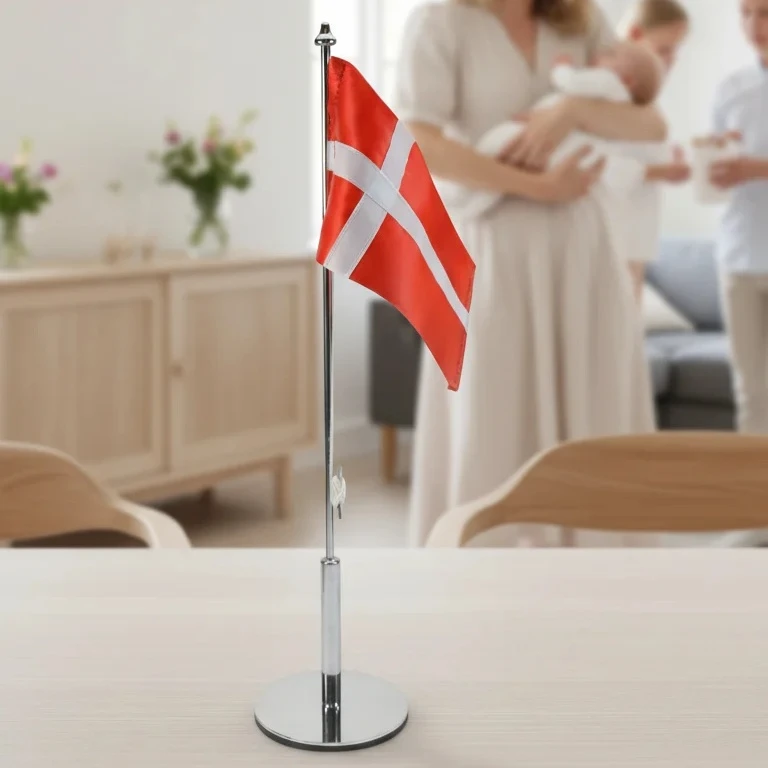 dansk-bordflag-dannebrog-med-metalstang Et dansk bordflag på en metalstang stående på et lyst træbord med en sløret familie i baggrunden til en festlig begivenhed.