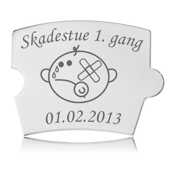 Memozz Spin brik i håndpoleret rustfrit stål med gravering af et grædende barn med plaster og teksten "Skadestue 1. gang" og en dato.