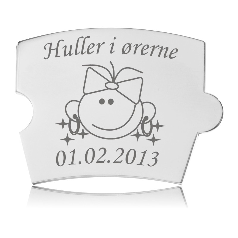 Memozz Spin mindebrik i håndpoleret stål med "Huller i ørerne" og en tegning af et smilende ansigt med sløjfe og øreringe.