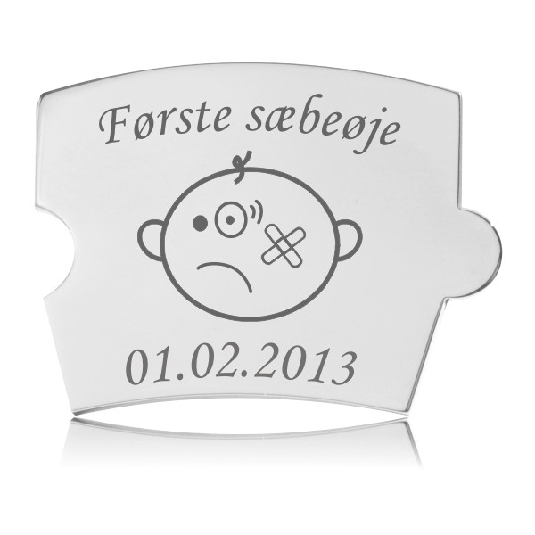 Memozz Spin brik i poleret rustfrit stål med teksten "Første sæbeøje" og tegning af barneansigt med forbinding